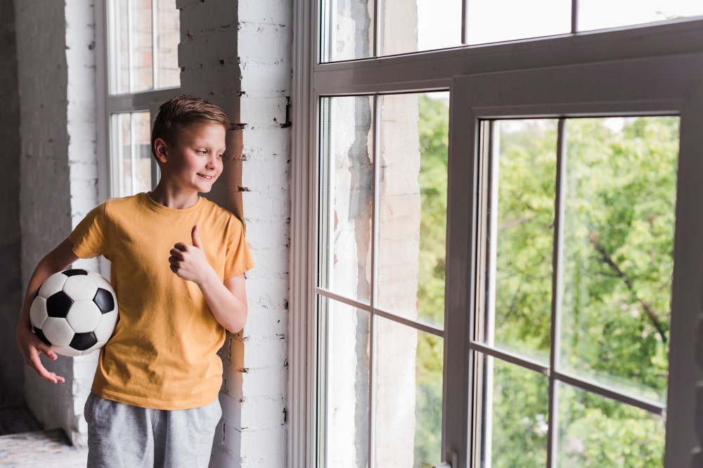 smiling-boy-holding-soccer-ball-looking-out-through-window-min